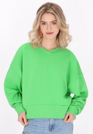 Grön överdimensionerad sweatshirt med V-ringning och ribbad mudd. Har en mjuk textur och en avslappnad passform, tillsammans med ljusblå jeans.