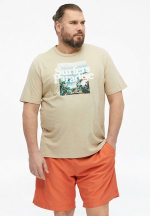 Mężczyzna z brodą i związanymi do tyłu włosami, ubrany w beżowy t-shirt "Surfers Paradise" i pomarańczowe szorty, stoi z jedną ręką w kieszeni, patrząc w prawo.