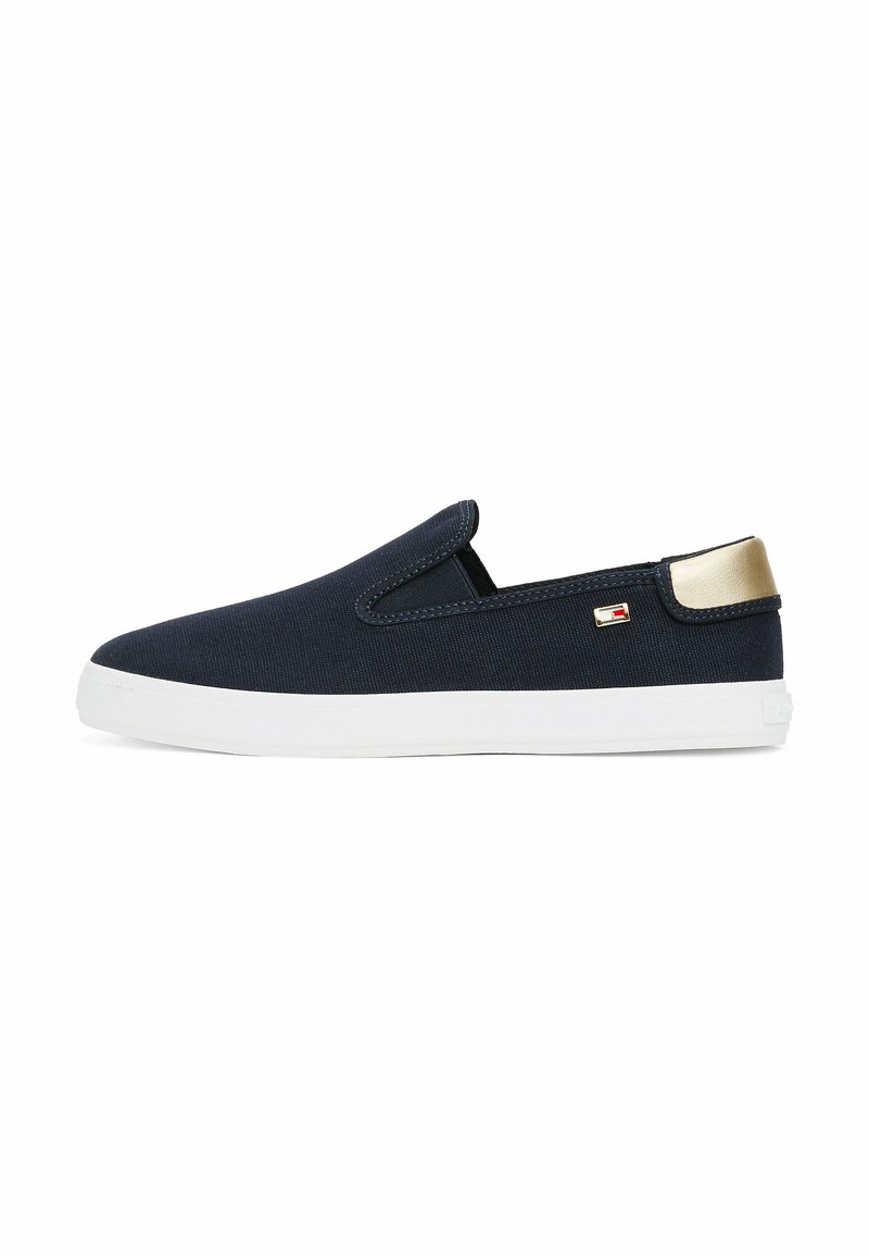 Scarpe slip-on navy con tomaia in tela testurizzata, suola in gomma bianca e un accento in metallo dorato sul tallone. Piccolo logo del marchio sul lato.