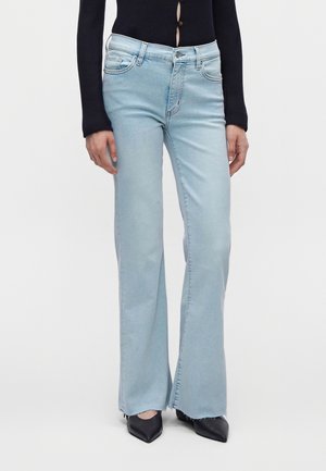 Personne portant un jean évasé bleu clair, des chaussures noires à bout pointu et un haut noir à manches longues avec des boutons, debout devant un fond uni.