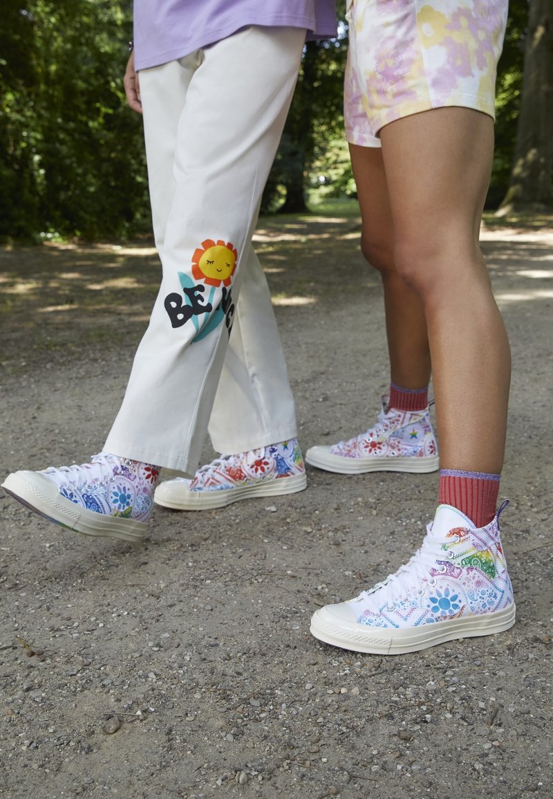 Deux personnes debout à l'extérieur portant des baskets montantes colorées avec des motifs floraux ; l'une porte un pantalon blanc avec une fleur souriante et un motif de texte.