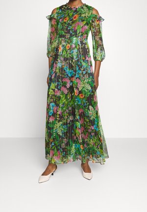 Robe longue fleurie avec des fleurs vertes, roses, bleues et oranges, découpes épaules dénudées, manches bouffantes transparentes, portée avec des chaussures blanches pointues à talons bas.