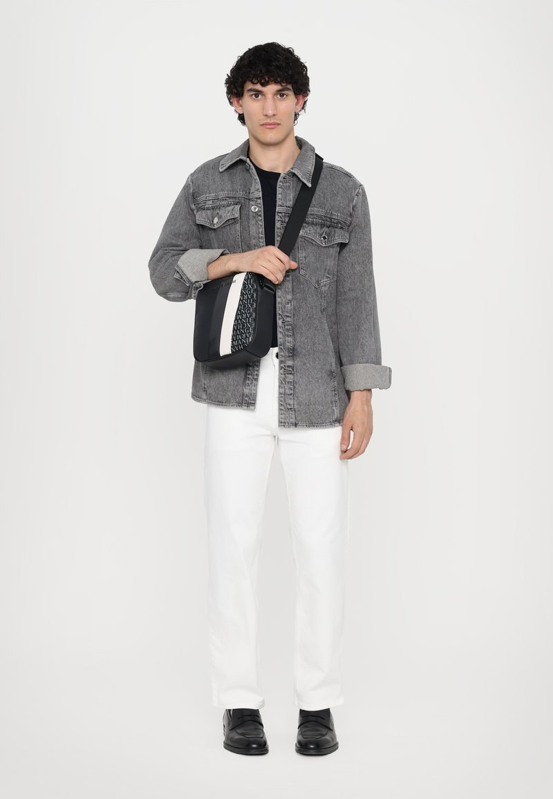 Svart og hvit skulderveske med et teksturert mønster, laget av syntetisk materiale, båret av en person i en grå denimjakke og hvite bukser.