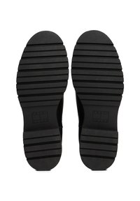 Semelles extérieures de chaussures noires avec de profondes rainures horizontales, présentant le logo Tommy Jeans en relief près de la zone du talon.
