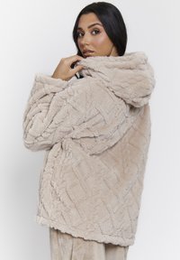 Beige, pluche hooded jack met een gestructureerd, verhoogd patroon. De stof lijkt zacht en comfortabel, met een ontspannen pasvorm en brede mouwen.