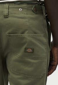 Άτομο με σκουρόχρωμο δέρμα που φοράει παντελόνι Dickies σε ελιά χρώμα, δείχνοντας τη μεγάλη πίσω τσέπη με το λογότυπο και μέρος ριγωτού πουκαμίσου πάνω από τη ζώνη.