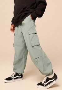 Hellgrüne Cargo-Hosen mit mehreren Taschen und elastischen Bündchen, kombiniert mit schwarzen Sneakers mit weißen Akzenten. Lässiger, entspannter Schnitt.