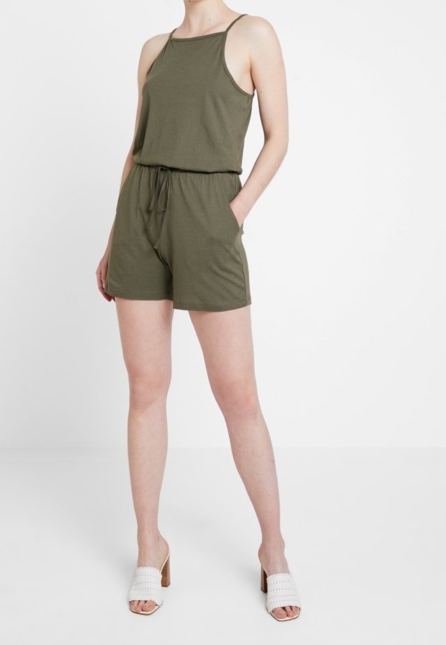 Olivgrön ärmlös jumpsuit med dragsko i midjan och sidofickor, i kombination med vita öppna klackskor med vävd design.