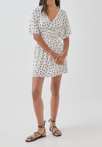 Valkoisessa pilkullisessa mekossa on V-pääntie, joustava vyötärö ja lyhyet hihat; yhdistettynä mustiin remmisandaaleihin. Yksinkertainen, arkinen muotoilu.