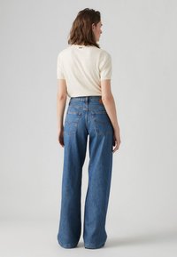 Aukštai juosmeni, plačios džinsinės kelnės, mėlynos spalvos, su kontrastinga siūle, užpakalinėmis kišenėmis ir ruda odine prekių ženklo etikete juosmens linijoje.