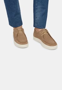 Pantofi din suede maro cu vârf rotund, talpă din cauciuc alb, și șireturi bej, asortați cu blugi albaștri desfăcuți.