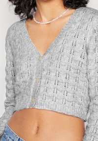 Szara krótka kardigan z wzorem dzianinowym i otwartym splotem oraz pięcioma kontrastującymi guzikami z przodu. Miękka struktura, ściągacz u dołu.