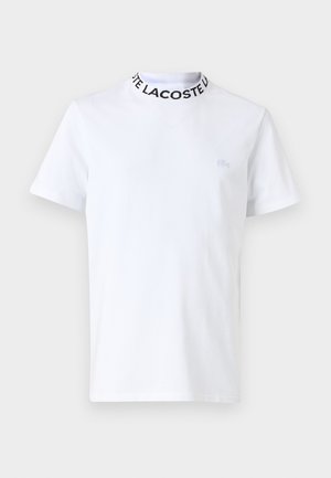 Bela majica iz bombaža, z okroglim črnim ovratnikom z besedilom "LACOSTE" in majhnim vezenim logotipom na prsih. Kratke rokave.