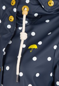 Imperméable bleu marine à pois avec boutons jaunes, logo parapluie jaune et cordon blanc avec détail perle en bois.