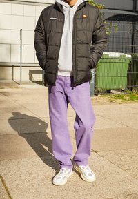 Czarna kurtka puchowa na szarym hoodie, zestawiona z jasnofioletowymi dżinsami. Obuwie ma białe i szare detale. Sceneria na zewnątrz z chodnikiem.