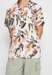 Chemise à manches courtes avec boutons, en tissu blanc orné de motifs floraux colorés et d'oiseaux tropicaux en vert, orange, rose et noir.