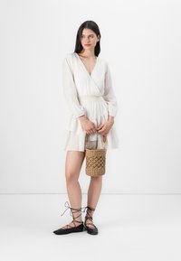 Una donna posa indossando un vestito avvolgente bianco e delle scarpe flat nere con lacci, mentre tiene una borsa intrecciata. Lo sfondo è minimalista e bianco.