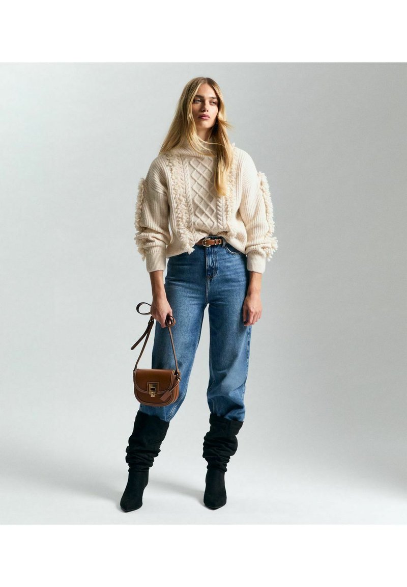 Suéter de punto crema con detalles texturizados, jeans azul de tiro alto, botas holgadas negras y un bolso bandolera marrón.