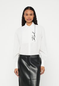 Camicia bianca con bottoni e colletto strutturato, con firma nera scritta a mano. Abbinata a una gonna in pelle nera a vita alta.