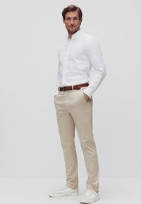 Witte button-up shirt, beige chinos en witte sneakers. De outfit heeft een gevlochten bruine riem, met een getailleerd ontwerp en een gladde textuur.