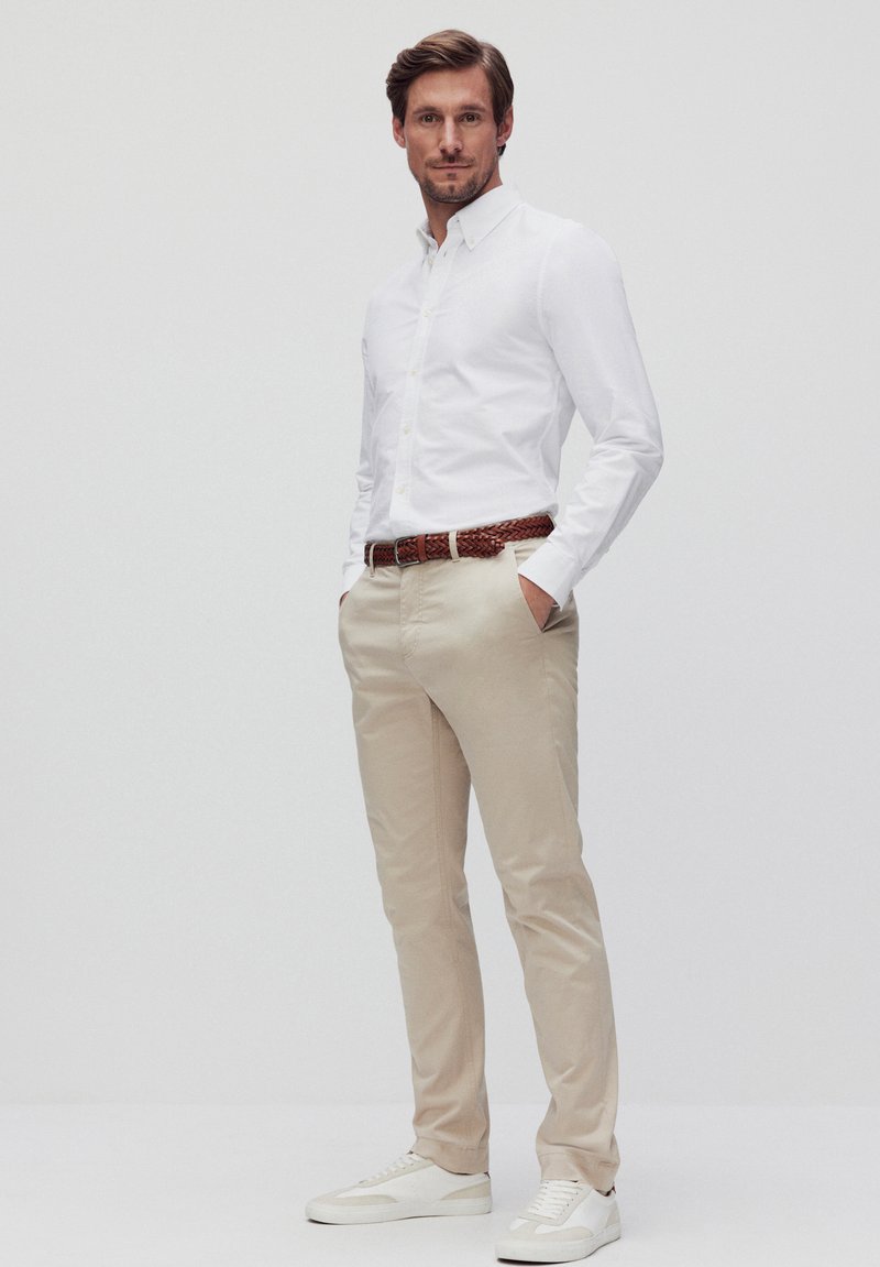 Witte button-up shirt, beige chinos en witte sneakers. De outfit heeft een gevlochten bruine riem, met een getailleerd ontwerp en een gladde textuur.