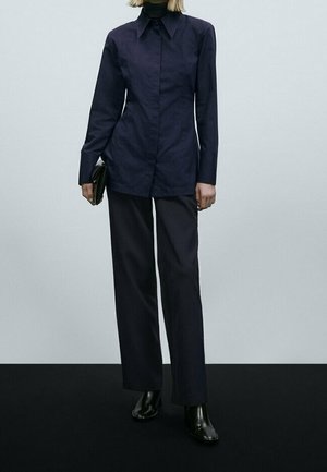 Camicia a maniche lunghe blu navy con colletto a punta, abbinata a pantaloni larghi scuri. Tessuto liscio e vestibilità sartoriale. Accessorio clutch nero.