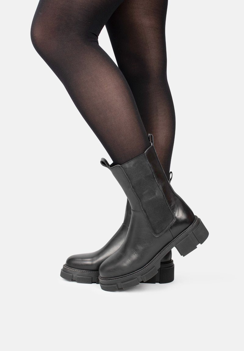 Botas de tobillo de cuero negro con paneles laterales elásticos, suelas de goma gruesas y tiradores. Textura suave y diseño de punta redonda.