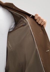 Braun gefütterte Jacke mit glatter Textur, ausgestattet mit einer Reißverschlusstasche und weißen Paspeln an den Kanten. Innenfutter in einem dunkleren Braun.