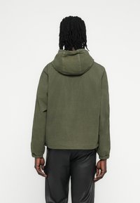 Ελιά πράσινο μπουφάν με κουκούλα και χαλαρή εφαρμογή, με ελαστικά μανίκια και λεία υφή. Απλός σχεδιασμός, χωρίς ορατά σχέδια ή διακοσμητικά στοιχεία.