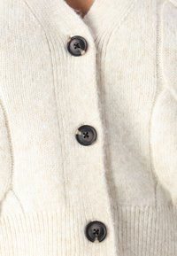 Cardigan de cor creme feito de lã macia, apresentando nervuras grossas, decote em V profundo e quatro grandes botões escuros na frente.