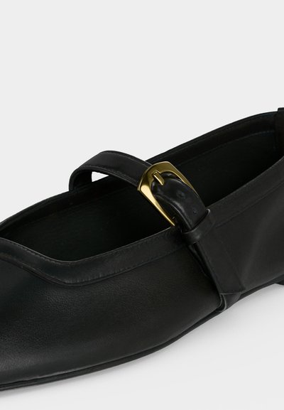 Chaussure plate noire en cuir avec une fine bride sur le dessus, fermée par une petite boucle dorée, sur un fond clair.