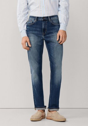 Homme portant un jean bleu délavé avec les revers retroussés, une chemise à rayures bleu clair et des espadrilles beiges sans lacets, debout devant un fond uni.