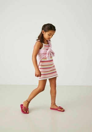 Gestreepte mouwloze jurk in roze en witte tinten, gecombineerd met een roze trui die om de schouders is geknoopt en roze slippers.
