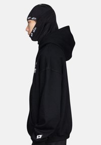 Haut à capuche noir avec cordon de serrage, arborant un logo sur le devant et une finition texturée. Le porteur porte un masque facial noir et un design avec capuche.