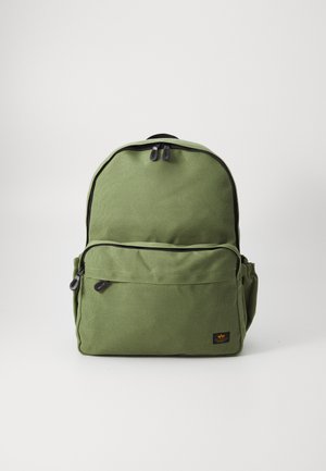 Mochila de lona verde con forma redondeada, cierre de doble cremallera, bolsillo frontal con cremallera, bolsillo lateral y un parche tejido de marca en la parte frontal inferior.