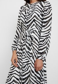 Svartvit klänning med zebramönster av lättviktigt material, långärmad, med knappstängning och ett knytbälte i midjan.