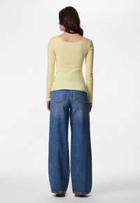 Kvinde med langt brunt hår iført en tætsiddende gul langærmet top og blå vidde-bukser, stående med ryggen mod på ensfarvet baggrund.