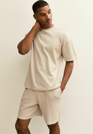 Man in beige korte mouwen shirt en bijpassende korte broek staat tegen een effen muur, met de ene hand aan zijn nek en de andere in zijn zak, kijkt naar beneden.