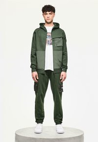 Groene hoodie met een voorzak, gecombineerd met bijpassende joggers met cargozakken. Gemaakt van zachte stof; gedragen met een wit graphic t-shirt.