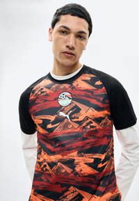 Νεαρός άνδρας με κοντά μαύρα μαλλιά φορώντας μαύρη και πορτοκαλί φανέλα ποδοσφαίρου της Αιγύπτου με γραφικά πυραμίδων και λογότυπο Puma.