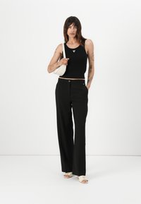 Svart avkortet tanktop med en trekantet detalj, kombinert med høyt midje sorte bukser. Modellen holder en liten hvit veske og har på seg åpne sko.
