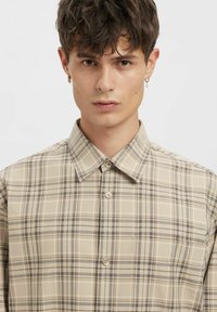 Beežkirju ruuduline särgipluus, millel on klassikaline krae, lühikesed varrukad ja heledapruunid nööbid, valmistatud siledast, tekstuuriga kangast.