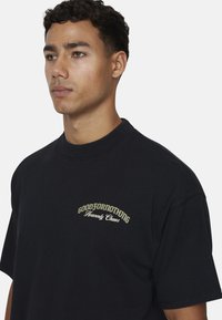 T-shirt in cotone nero con collo rotondo, maniche corte e ricamo frontale in oro con la scritta "GOODFORNOTHING Heavenly Chaos."