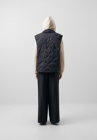 Chaleco acolchado sin mangas en un azul marino oscuro con un patrón de acolchado, llevado sobre una sudadera crema y combinado con pantalones sueltos oscuros. Vista desde atrás.