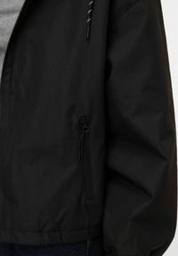 Sort jakke med lynlåslomme i siden og synligt snoretræk, båret over en grå skjorte og mørkeblå jeans.