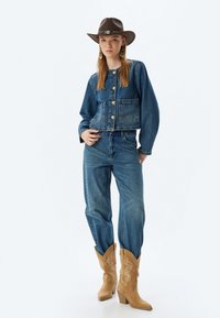 Chaqueta de denim con botones dorados y bolsillos delanteros, combinada con jeans de denim de tiro alto. Sombrero de cowboy marrón y botas de cowboy color cuero con detalles bordados.