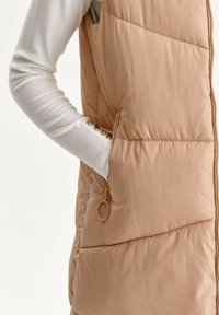 Beige Steppweste mit gestepptem Muster, ausgestattet mit einer Seitentasche und einem Reißverschluss mit goldenem Ring. Getragen über einem weißen Langarmoberteil.