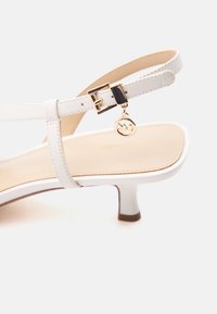 Λευκό δερμάτινο σανδάλι με χαμηλό τακούνι, λουρί στο αστράγαλο, χρυσή πόρπη και ένα μικρό στρογγυλό χρυσό φυλαχτό που κρέμεται από το λουρί.