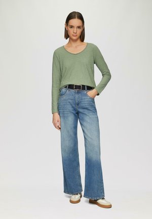Lange mouwen, lichtgroene top met een V-hals, gecombineerd met wijde blauwe denim jeans en beige sneakers met een textuurzool en zwarte accenten.