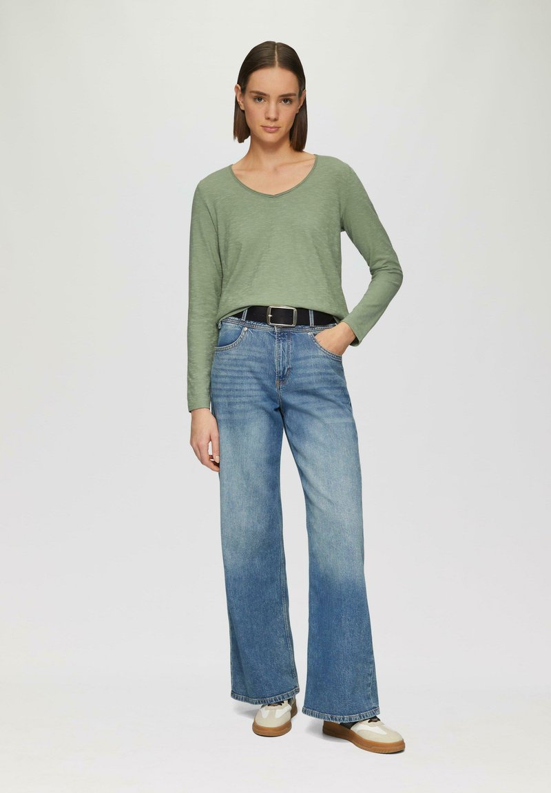 Lange mouwen, lichtgroene top met een V-hals, gecombineerd met wijde blauwe denim jeans en beige sneakers met een textuurzool en zwarte accenten.